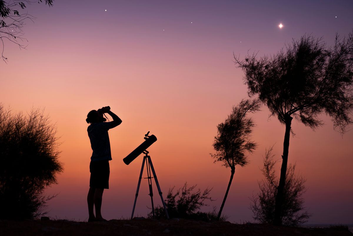 A man standing next to a portable telescope uses binoculars to observe stars in the twilight sky