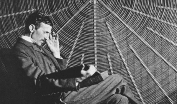 Black and white photograph of Nikola Tesla in front of the spiral coil of his high-voltage Tesla coil transformer