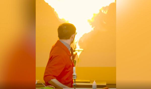 Science demonstrator in red lab coat faces away, looking at a plume of flames