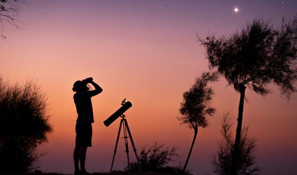 A man standing next to a portable telescope uses binoculars to observe stars in the twilight sky