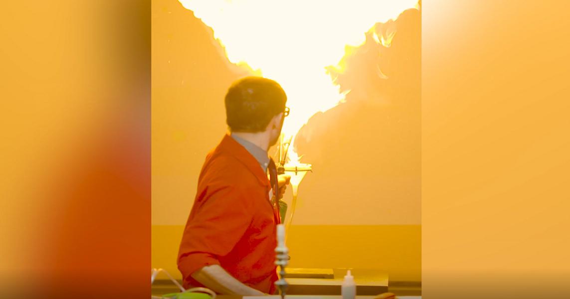Science demonstrator in red lab coat faces away, looking at a plume of flames